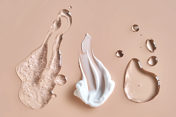 Texturproben flüssiger und halbfester Kosmetik: klares Serum, weiße Creme und Tropfen auf beigem Hintergrund.