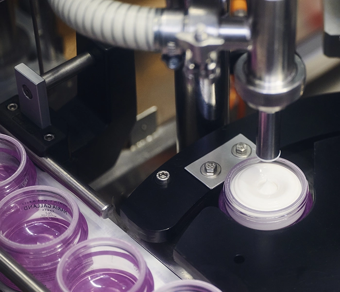 Abfüllanlage dosiert weiße Creme in einen Tiegel, violette Kosmetiktiegel auf dem Transportband.