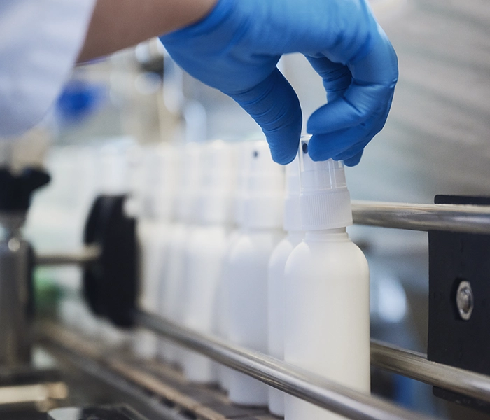 Hand in a blue glove places white pump spray bottles on the filling line for medical devices.