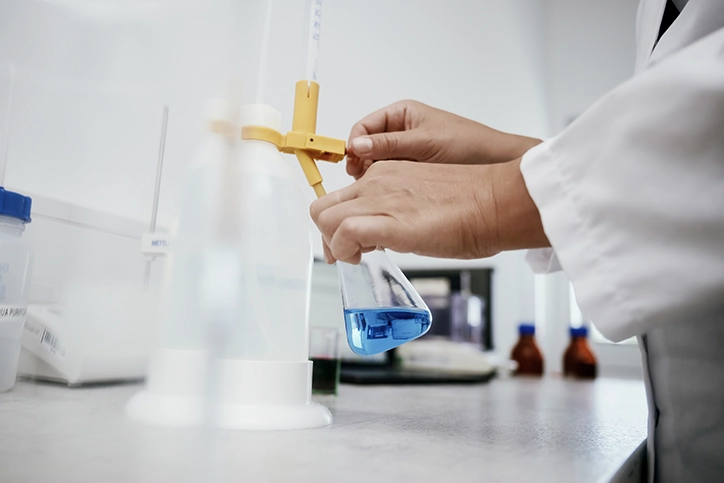 Labormitarbeiterin im weißen Kittel prüft eine blaue Rezeptur im Erlenmeyerkolben in der Qualitätskontrolle.